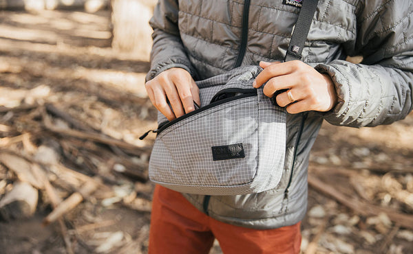 Person wearing the Design Lab Edition Packing Cube Shoulder Bag in Northwest Sky while standing in a forest. They are opening the front zipper pocket.